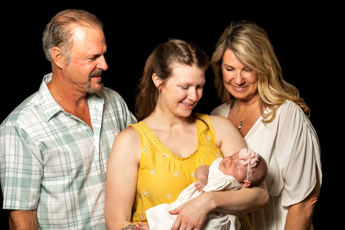 A family of four, including an older man, an older woman, a young woman, and a newborn girl, are gathered together and smiling against a black background. The young woman is holding the baby girl, who is wearing a white dress and a pink headband with