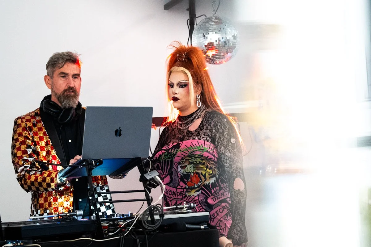 A DJ with gray hair and a beard wearing a checkered blazer stands next to a woman with long, bright orange hair styled in a high ponytail, wearing makeup and a colorful, graphic shirt. They are working on a laptop and DJ equipment, with a disco ball 