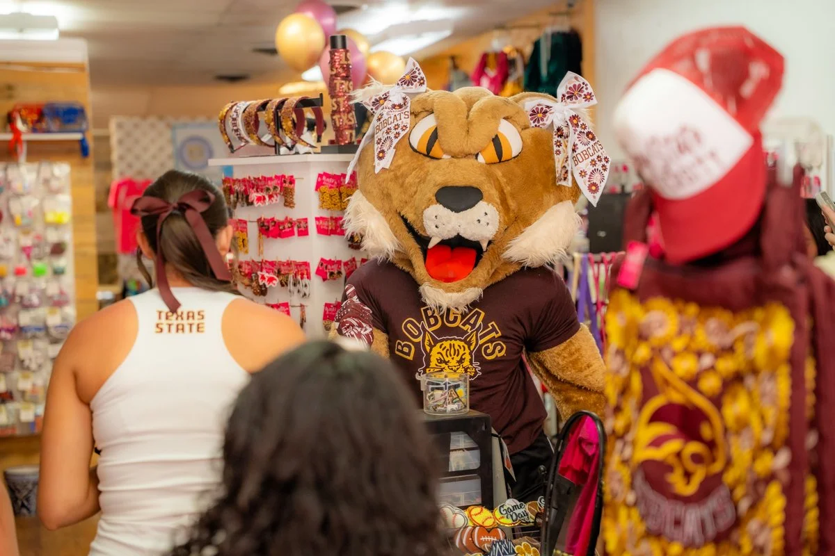 A person dressed in a Bobcats mascot costume with a lion's head, orange eyes, and a brown T-shirt saying 'BOBCATS'. They are at a store decorated with maroon and gold, with a woman in a Texas State shirt and other shoppers nearby.