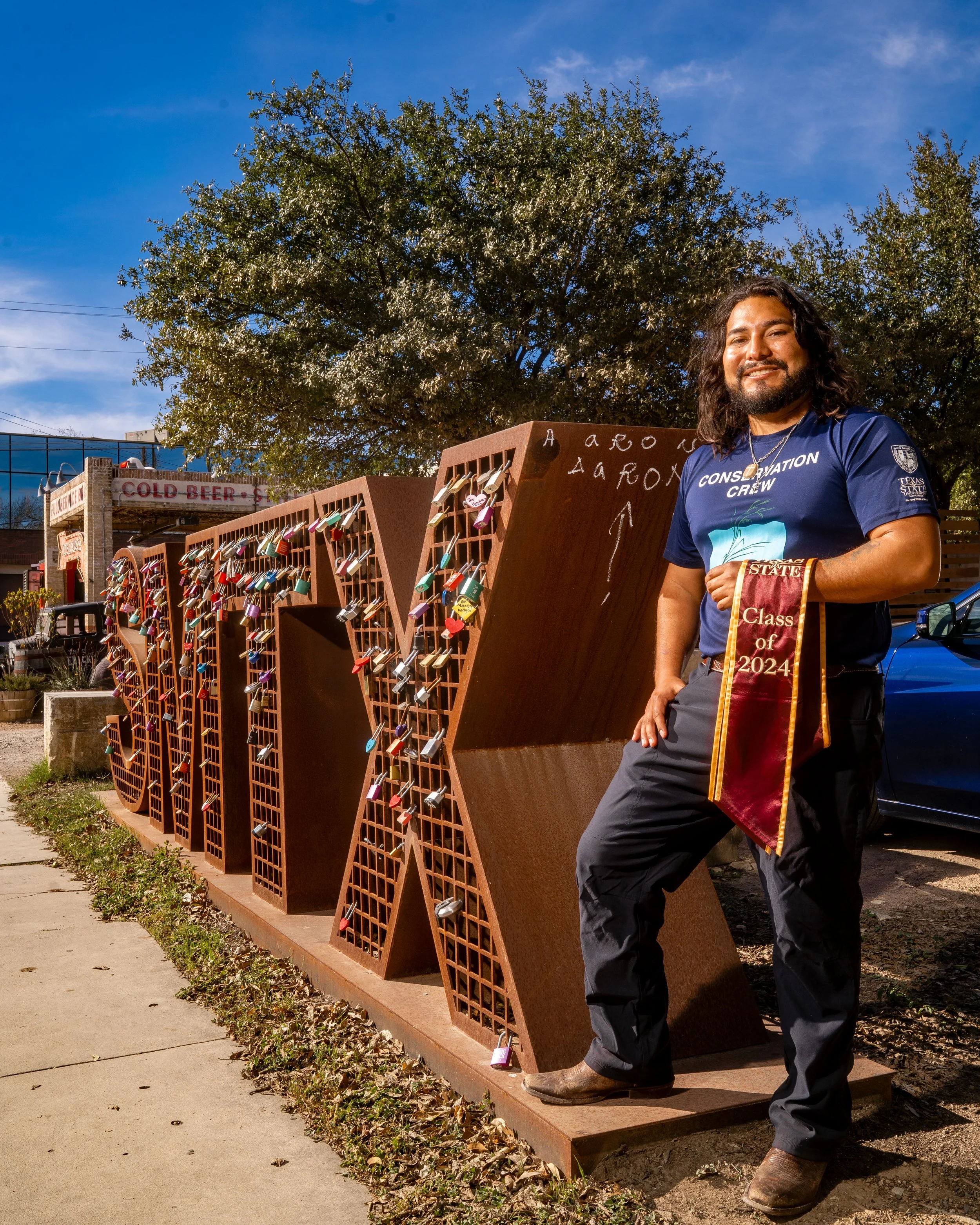 A man holding a maroon graduation stole with gold trim and 'Class of 2024' embroidered on it, standing next to a large metal sculpture of the word 'LOVE' decorated with numerous colorful padlocks, outside on a sunny day with a tree and a building in 