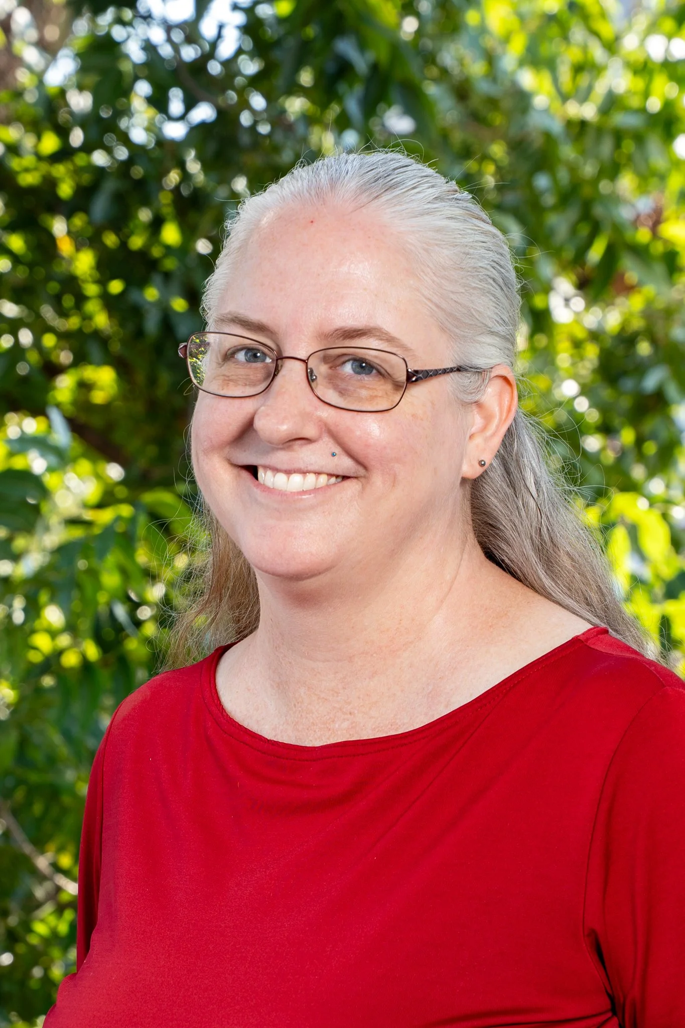 Smiling woman with gray hair, glasses, and a small nose piercing, wearing a red top, outdoors with leafy green background.