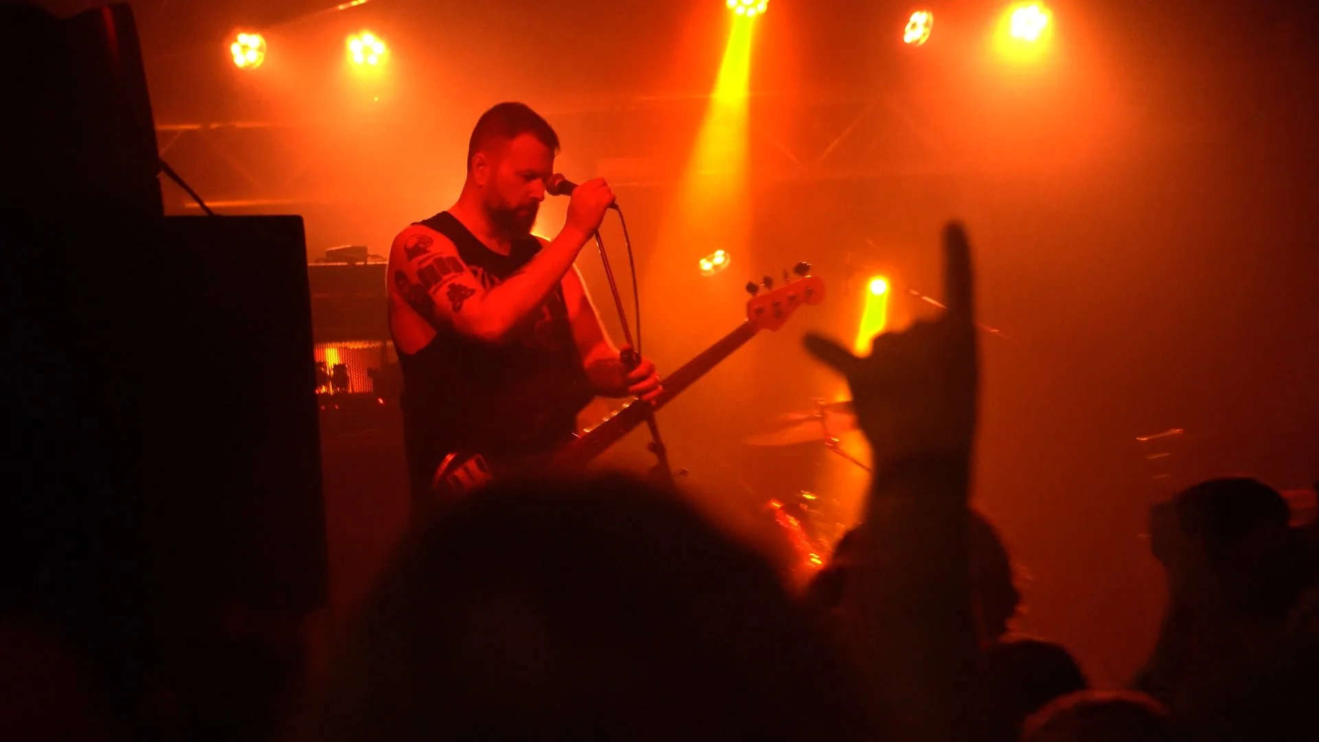 A musician with tattoos on his arms performs on stage with orange and yellow lighting, holding a microphone and playing a bass guitar.
