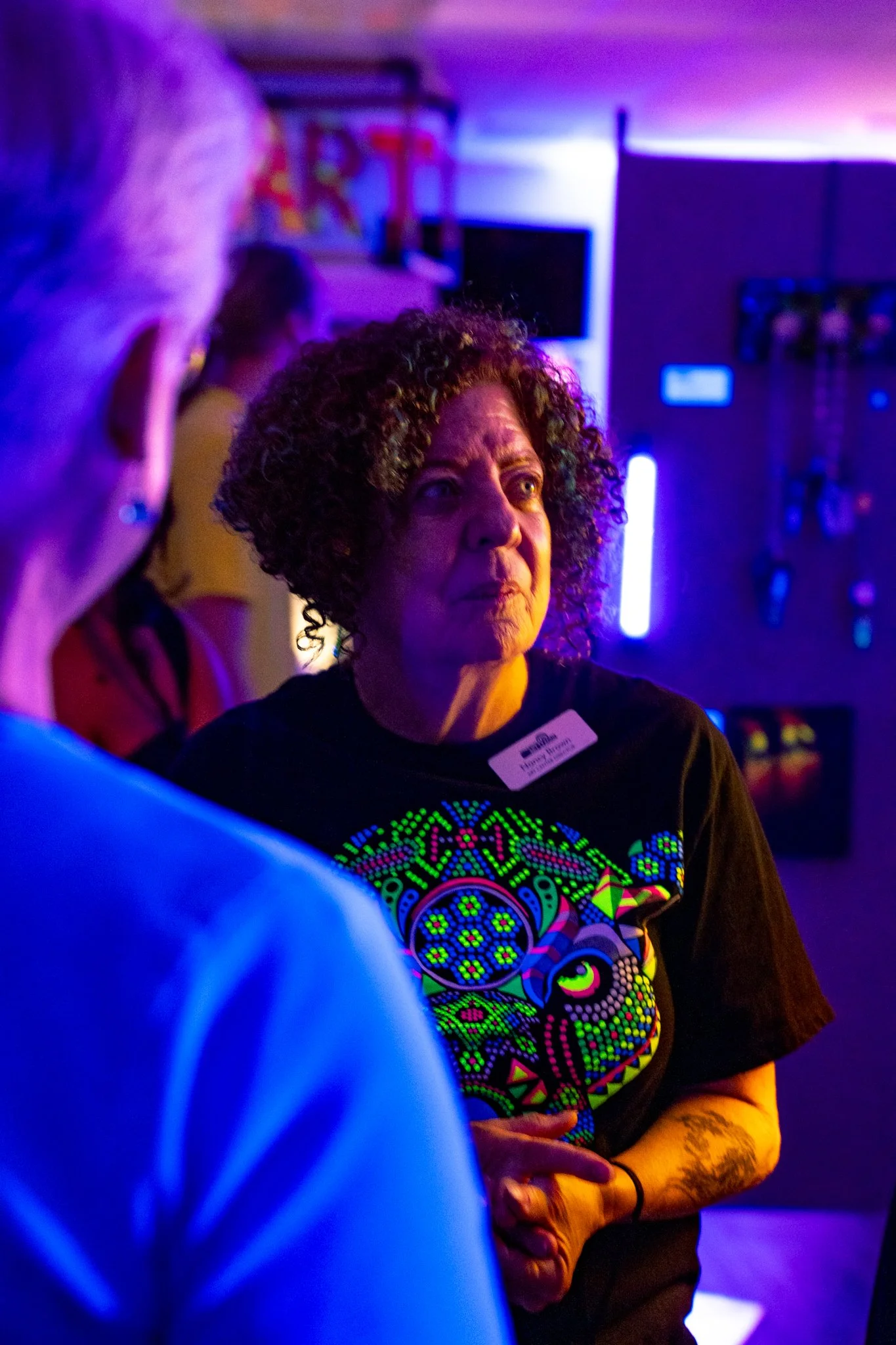 A woman with curly hair talking to another person in a room illuminated by vibrant blue and purple lighting, with artwork and tools displayed in the background.