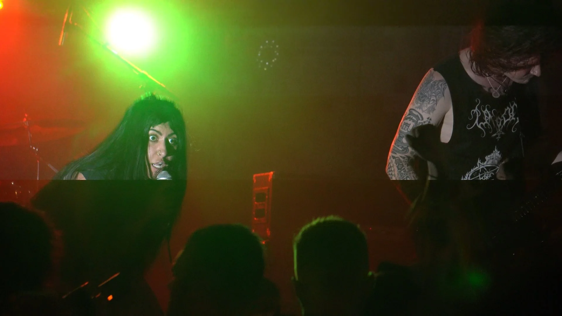 A woman with black hair and tattoos, singing into a microphone on stage with colorful lighting, alongside another person playing guitar.