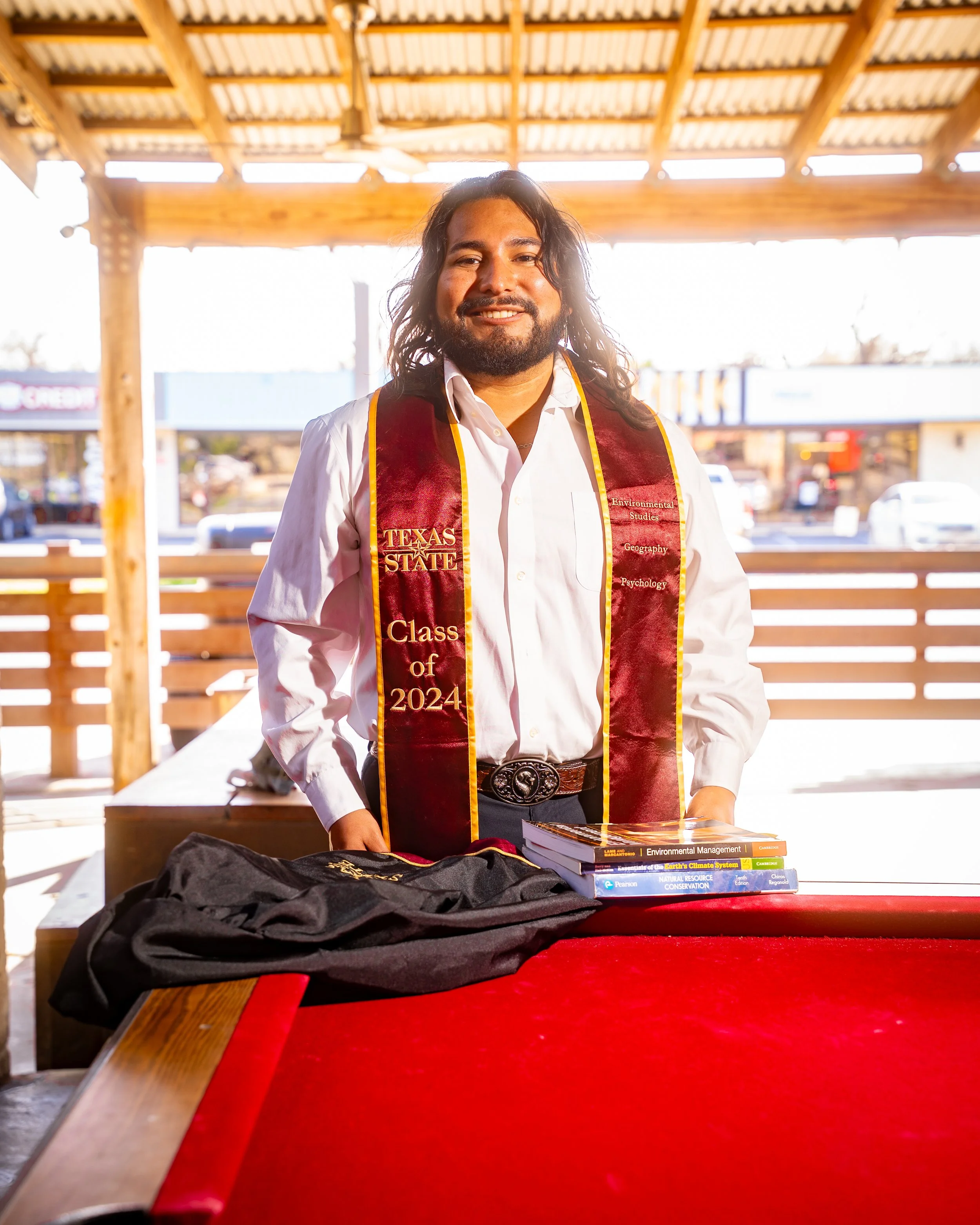 A man in a white shirt and maroon graduation stole with gold trim, reading 'Texas State Class of 2024,' stands at a table with books and a black graduation gown, inside an outdoor wooden pavilion.