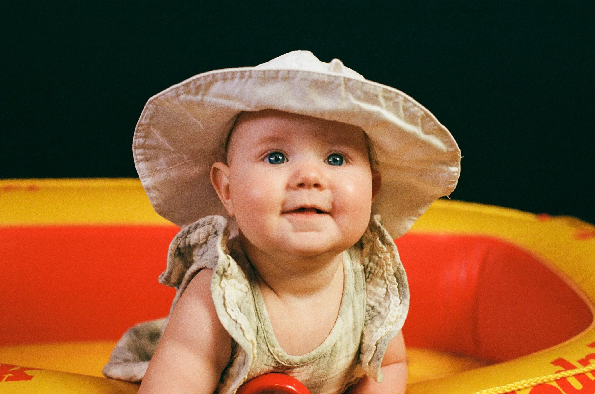 A baby with blue eyes wearing a beige sleeveless outfit and a wide-brimmed beige hat, lying on a yellow and red inflatable boat or pool float.