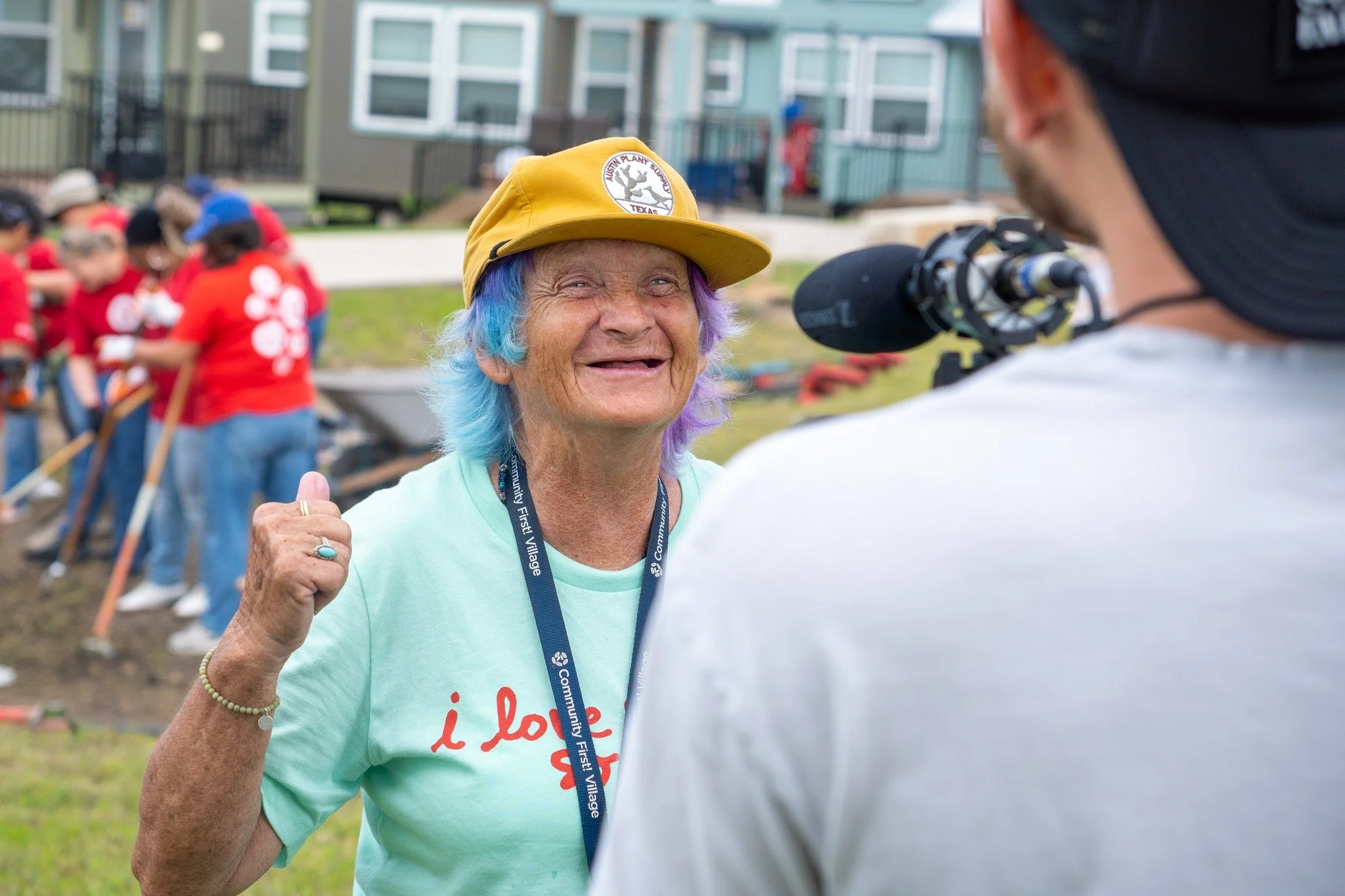 An elderly woman with multi-colored hair and a yellow cap smiling and talking to a person with a camera during a community event outdoors with volunteers in the background.