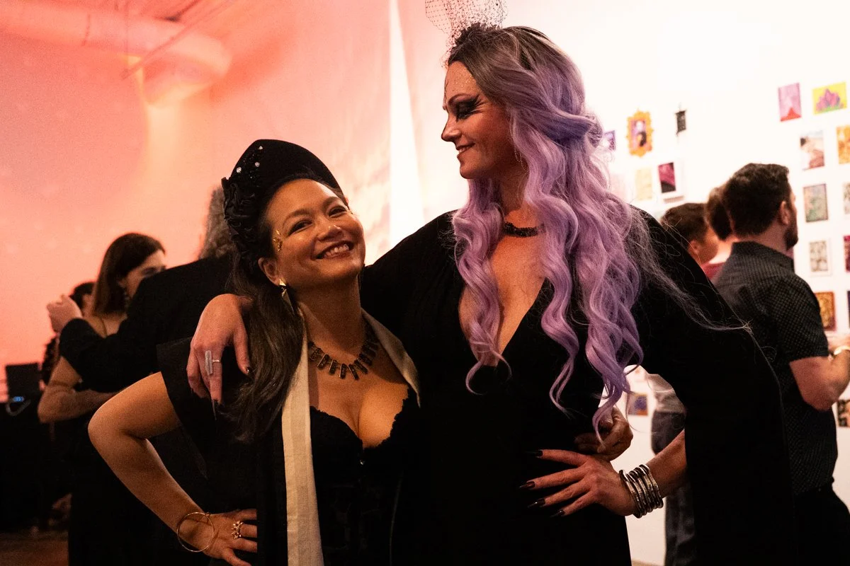 Two women at an art gallery, one with dark hair and the other with long lavender hair, smiling and posing together with their arms around each other.
