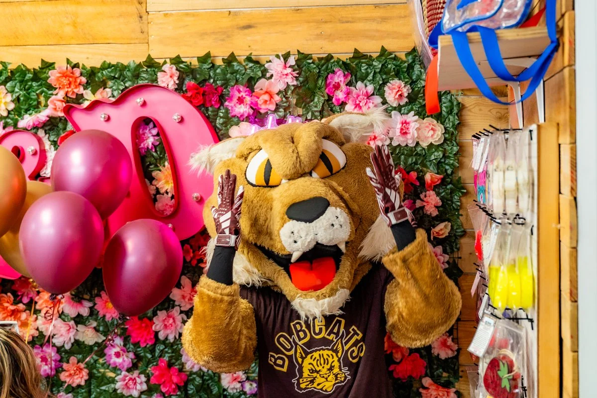 A lion mascot with a fuzzy face, wearing a black jersey that says "BOBCATS", holding its head with both paws, standing in front of a floral pink and green backdrop with balloons and decorations.