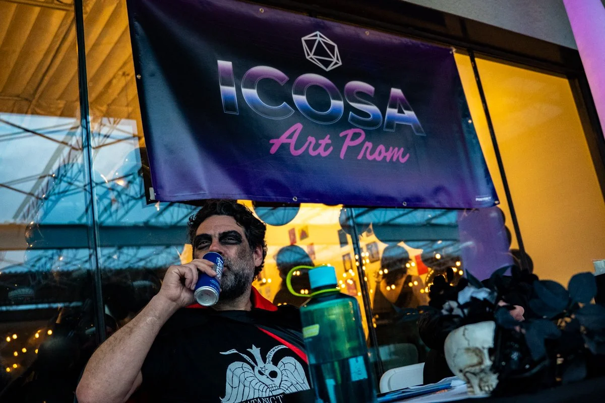 A man is sitting at a table drinking from a can at an event called I.C.O.S.A Art Prom, with a purple and pink banner hanging above him. The banner has the text "I.C.O.S.A Art Prom" and a geometric design. The background shows a glass window reflectin