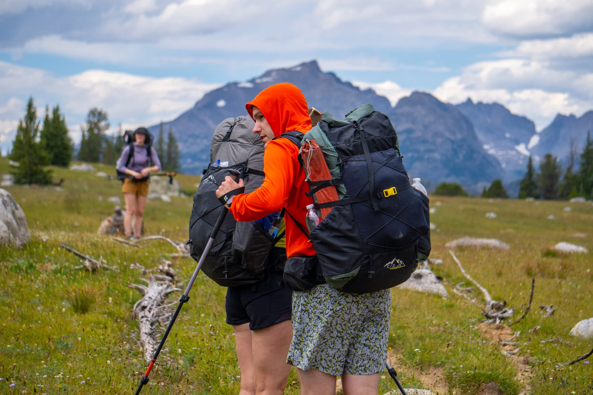 Two hikers, one in an orange hoodie and patterned shorts, and the other in a purple jacket and brown shorts, carrying large backpacks with mountains in the background.