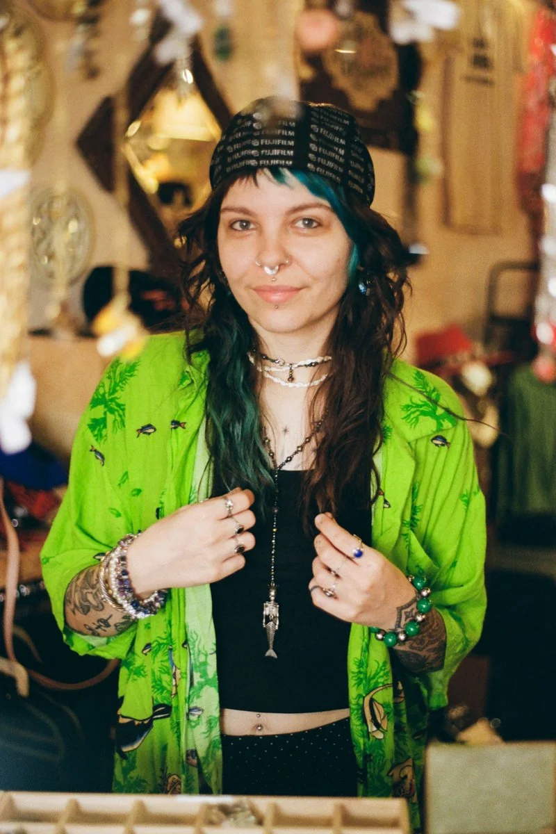 A woman with dark, wavy hair and teal streaks wearing a black and white striped bandana, multiple necklaces, rings, and bracelets. She is dressed in a black top and a vibrant green shirt with a pattern, standing indoors with a warm, decorative backgr