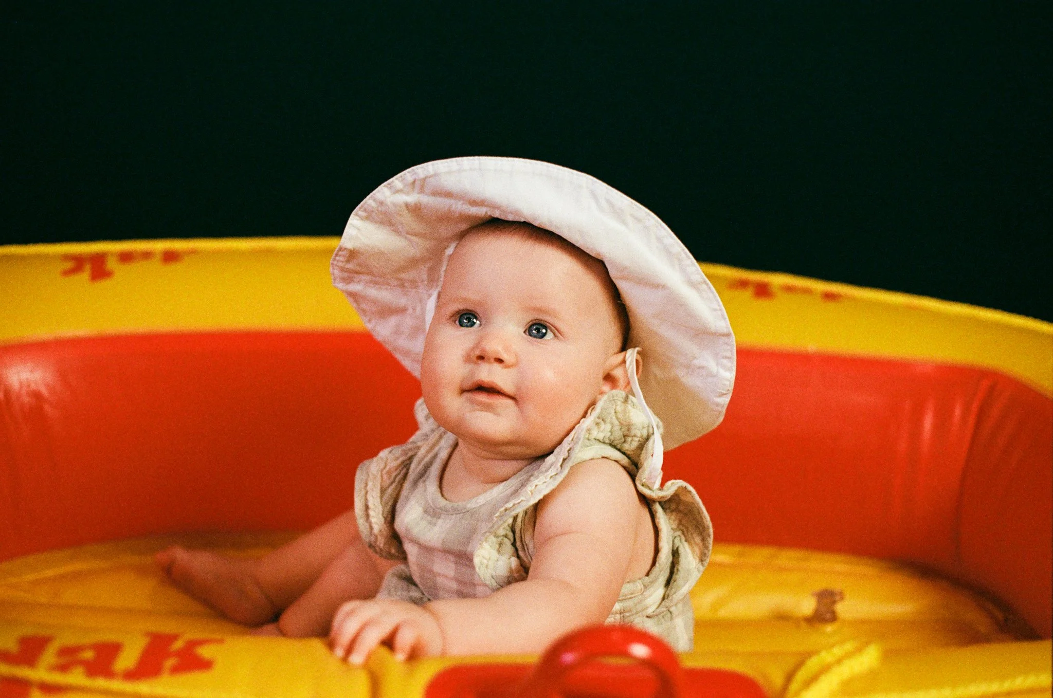 A baby wearing a sunhat sitting inside a colorful splash pad or water play area with yellow, red, and orange accents.