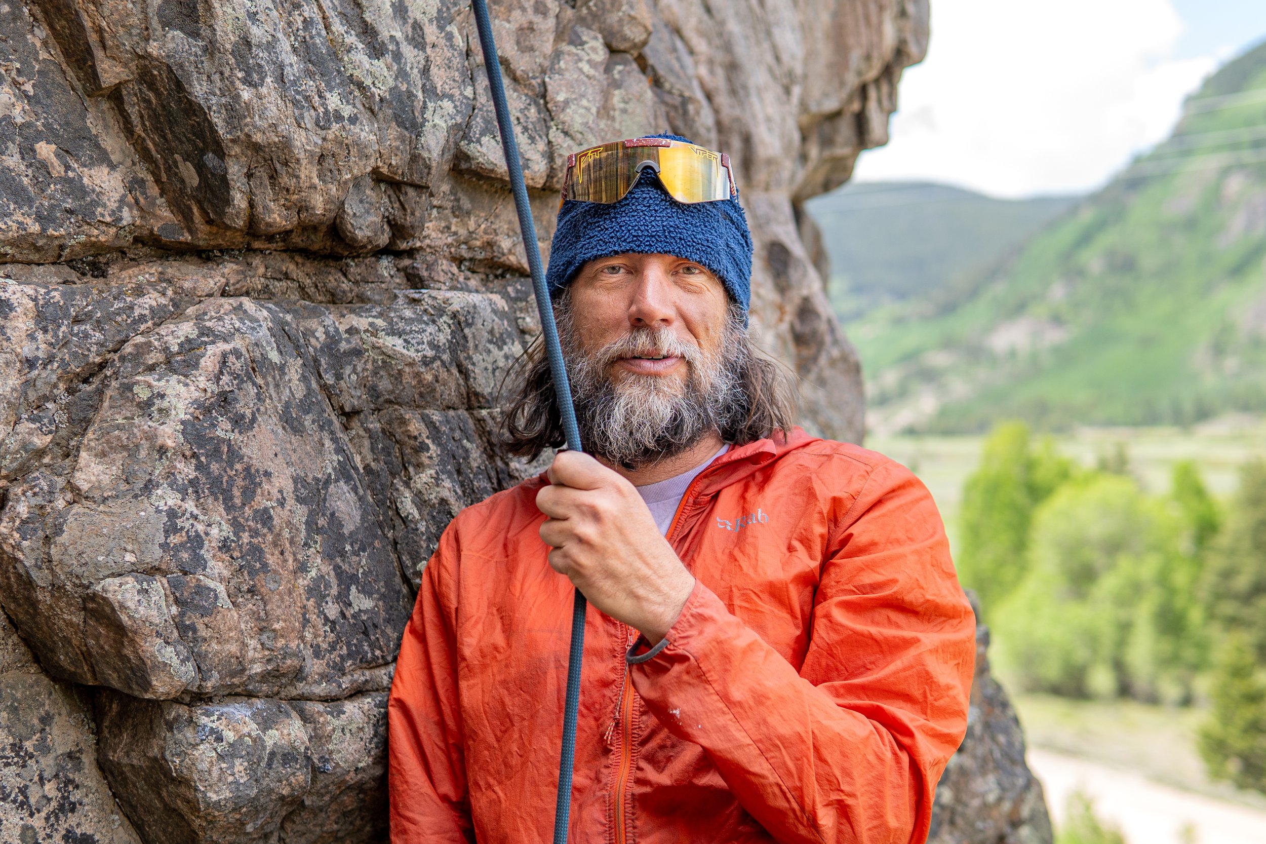 A man with a beard and long hair holding climbing gear, standing next to a rock face outdoors, wearing an orange jacket, a blue knit cap, and reflective sunglasses with a mountainous and green landscape in the background.