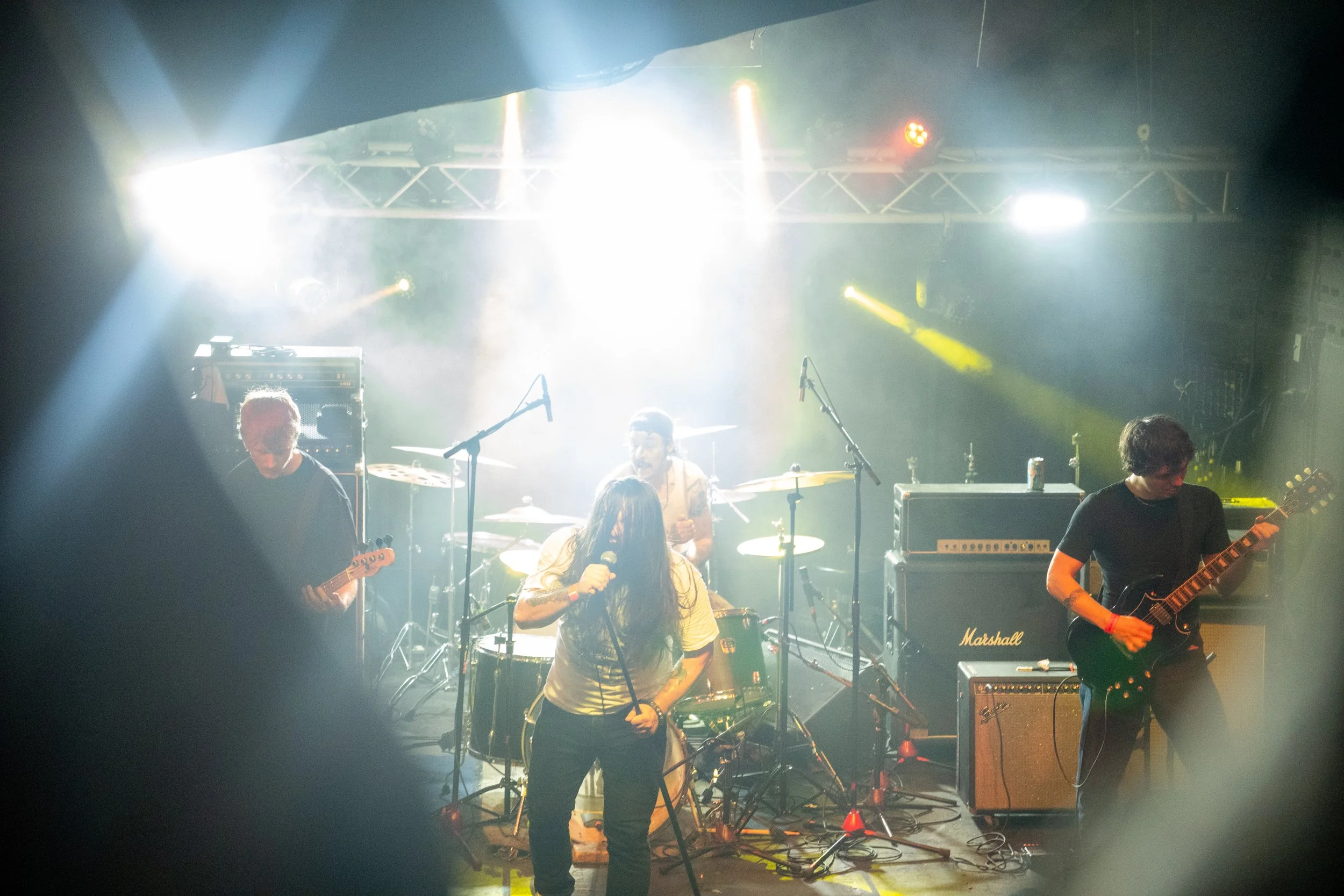 A band performing on stage with bright lights, including a female vocalist with long dark hair, a drummer, and two guitarists, one on the left and one on the right, with musical equipment around them.