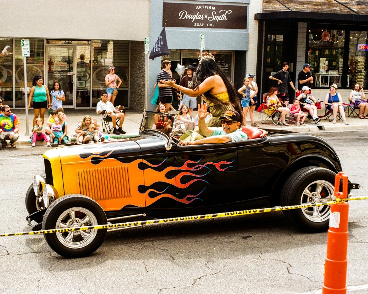 A vintage black car with flame decals on the side driving down a street during a parade, with a woman standing in the car handing out items to a smiling man waving from the passenger seat, while spectators sit and stand along the sidewalk watching.