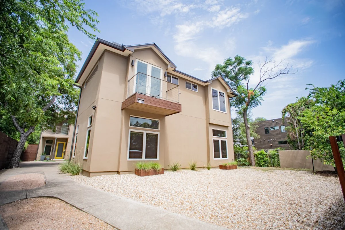 A modern, multi-story house with beige walls and white framing, featuring a small balcony; the yard is covered with gravel and has some plants, surrounded by trees and neighboring houses under a partly cloudy sky.