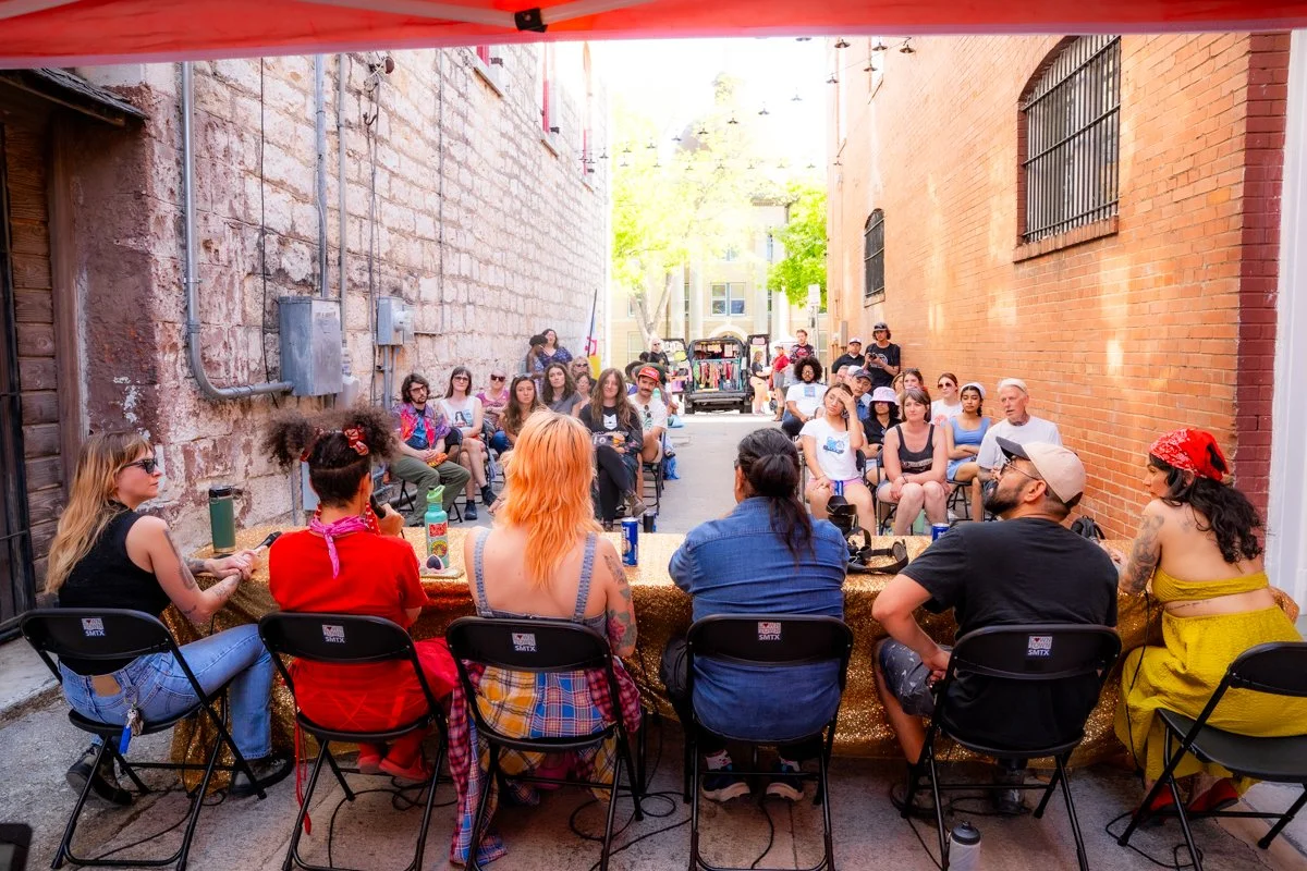 A panel of six people sitting at a table outdoors, addressing an audience of diverse individuals seated in a narrow alley between brick buildings, with some trees and a vehicle in the background.