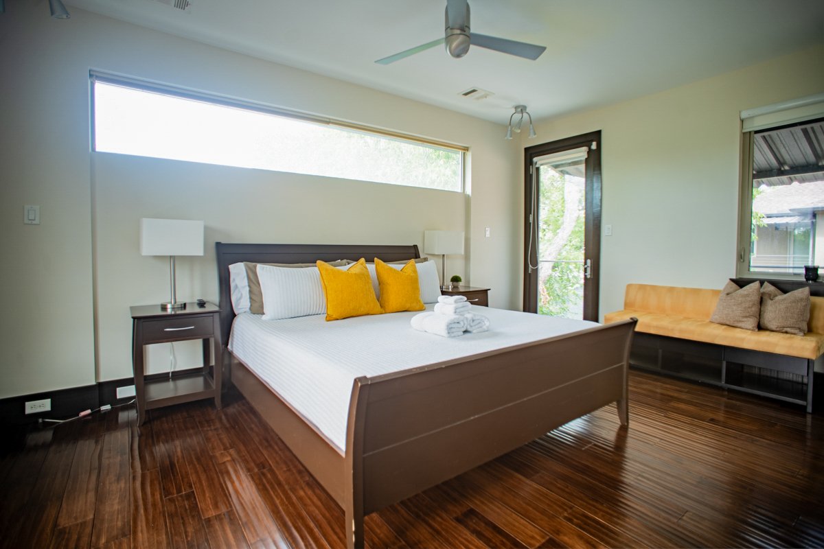 Modern bedroom with a bed, yellow pillows, white linens, wooden furniture, and windows with views of greenery.