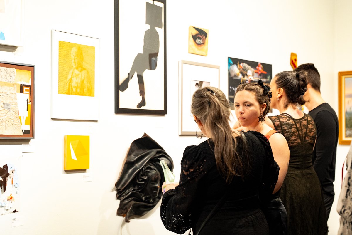 A group of people at an art gallery, looking at various framed artworks on the wall, with some engaging in conversation.