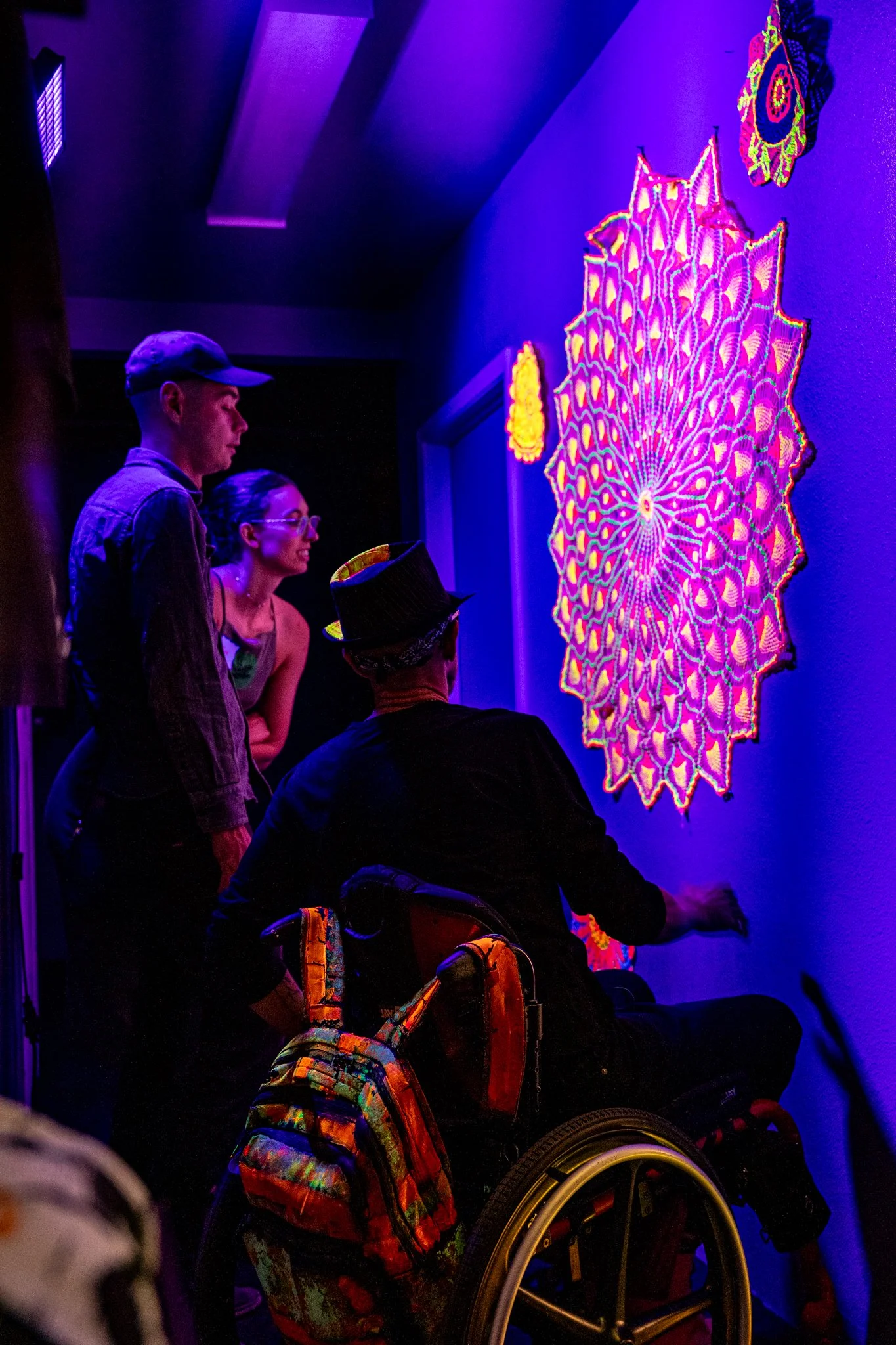 People observing glowing, colorful art pieces in a dark room, including one in a wheelchair.