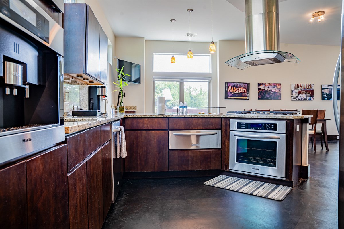 Modern kitchen with dark wooden cabinets, stainless steel appliances including an oven and a coffee machine, granite countertops, a potted plant, and colorful artwork on the wall, with a dining area in the background.