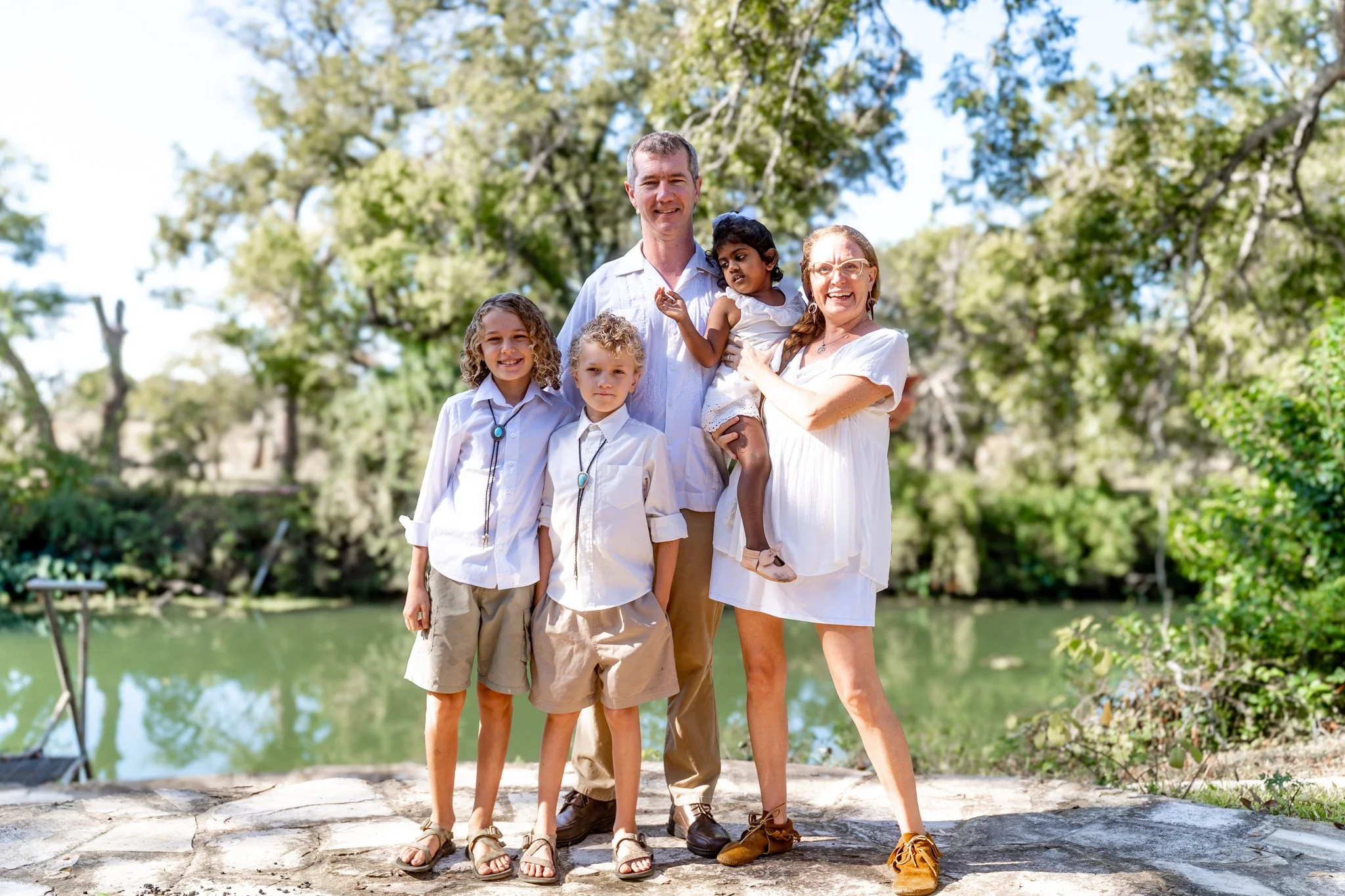 Family of six standing outdoors near a lake with trees in the background, smiling, wearing white and beige clothing.