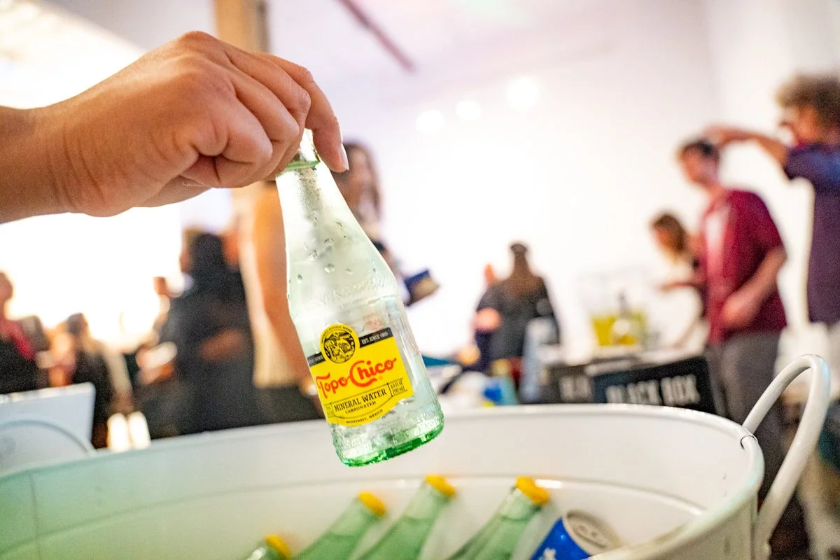 A hand dropping a Topo Chico sparkling mineral water bottle into a cooler at a crowded indoor event.
