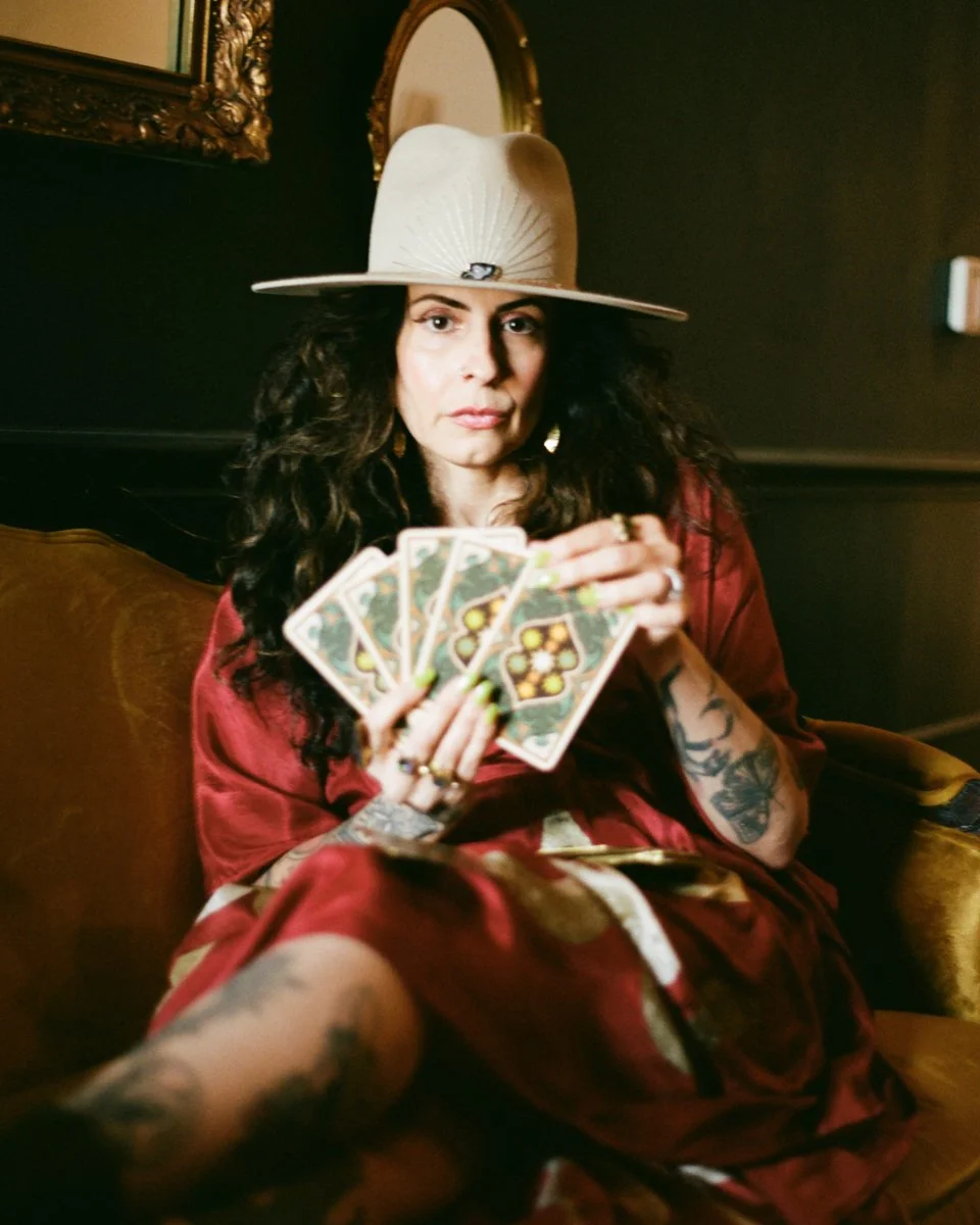 A woman with curly dark hair wearing a large beige hat, red dress, and fishnet stockings, sitting on a vintage-style yellow sofa, holding tarot cards.