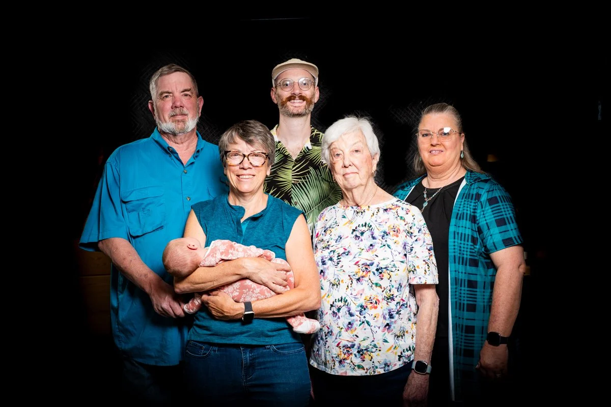 A group of six older adults, including a woman holding a newborn baby, standing against a black background.