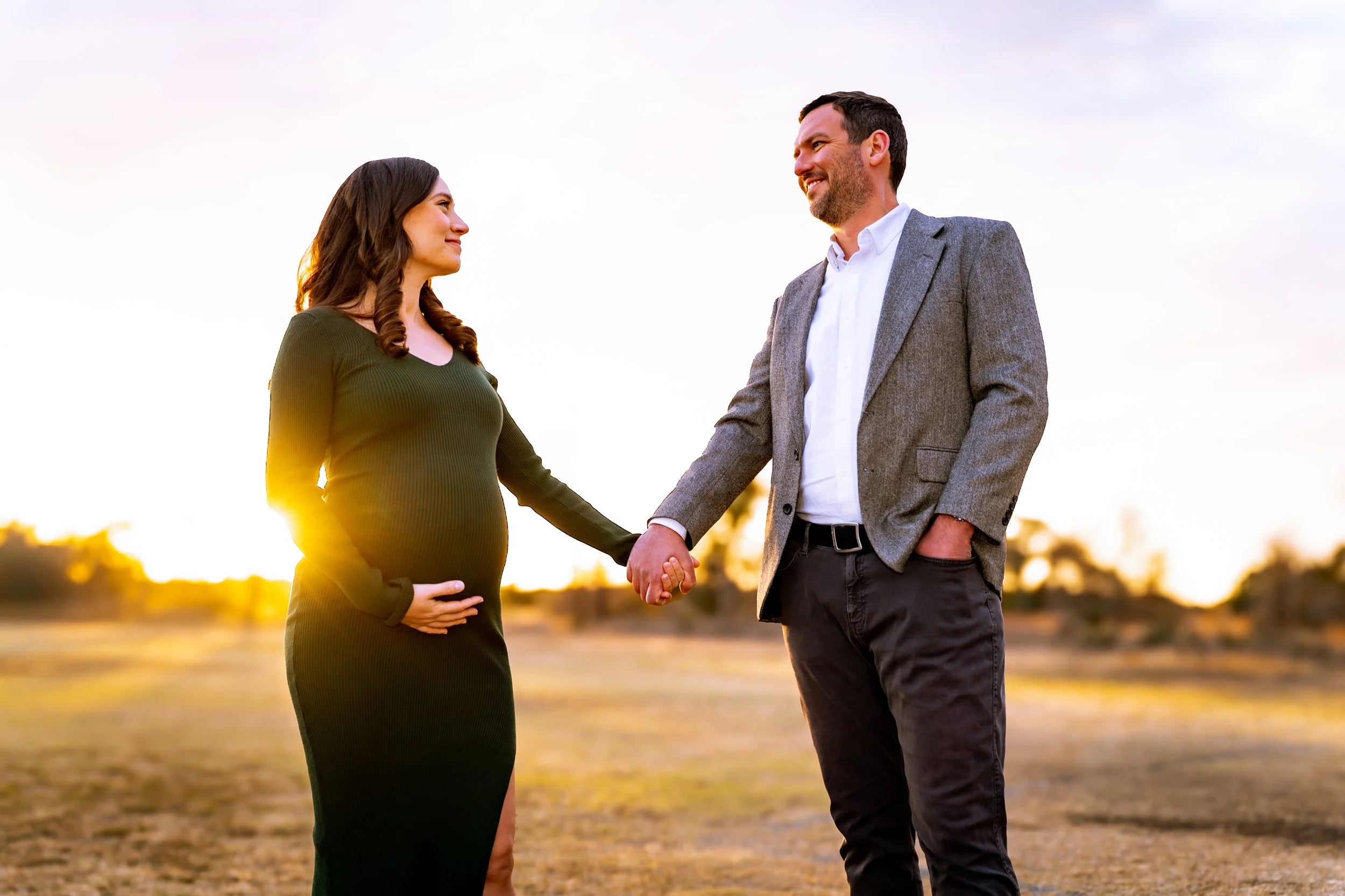 A pregnant woman standing outdoors at sunset, holding her belly with one hand, and holding hands with a man dressed in a blazer. They are facing each other with smiles, standing on a dirt field with blurred trees and sky in the background.