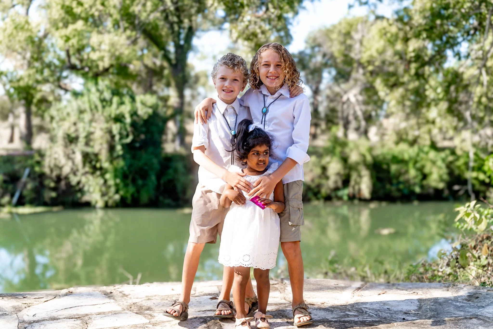 Three children, two girls and one boy, standing together outdoors near a lake, smiling at the camera with greenery in the background.