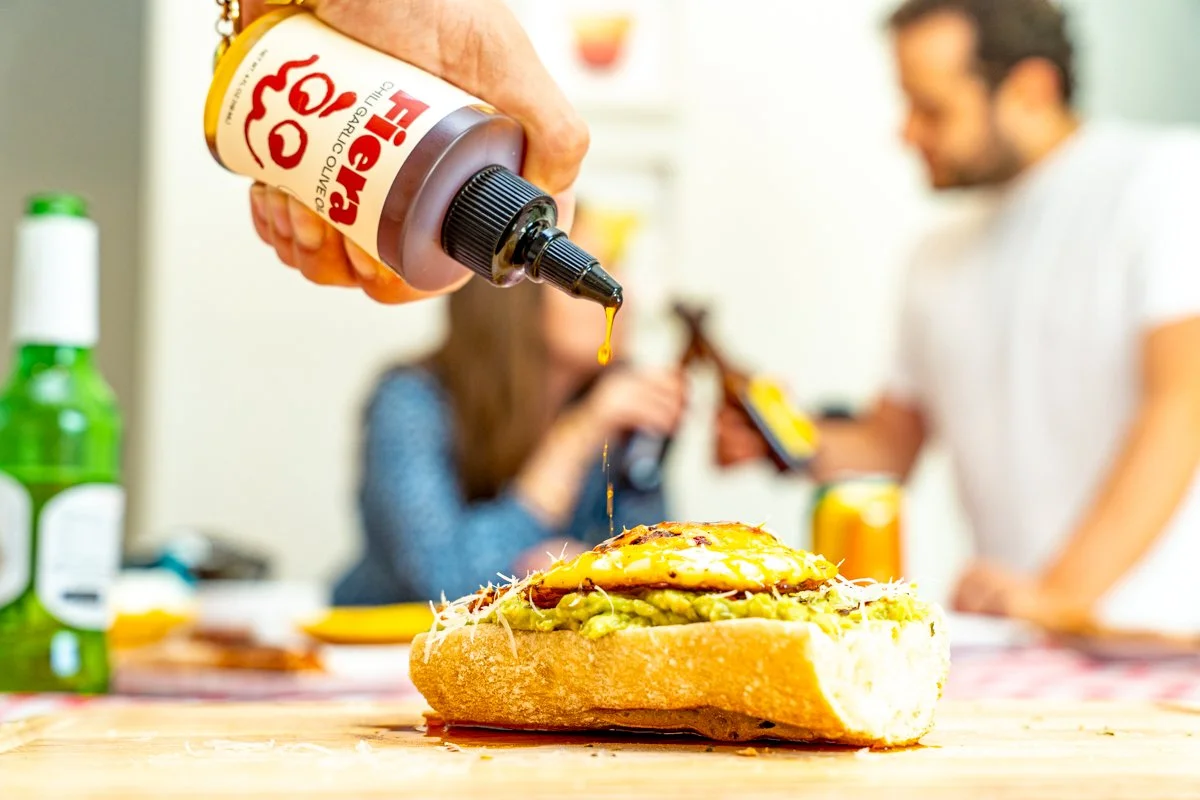 A sandwich on a wooden surface with mustard being squeezed from a bottle over it, with people in the background.