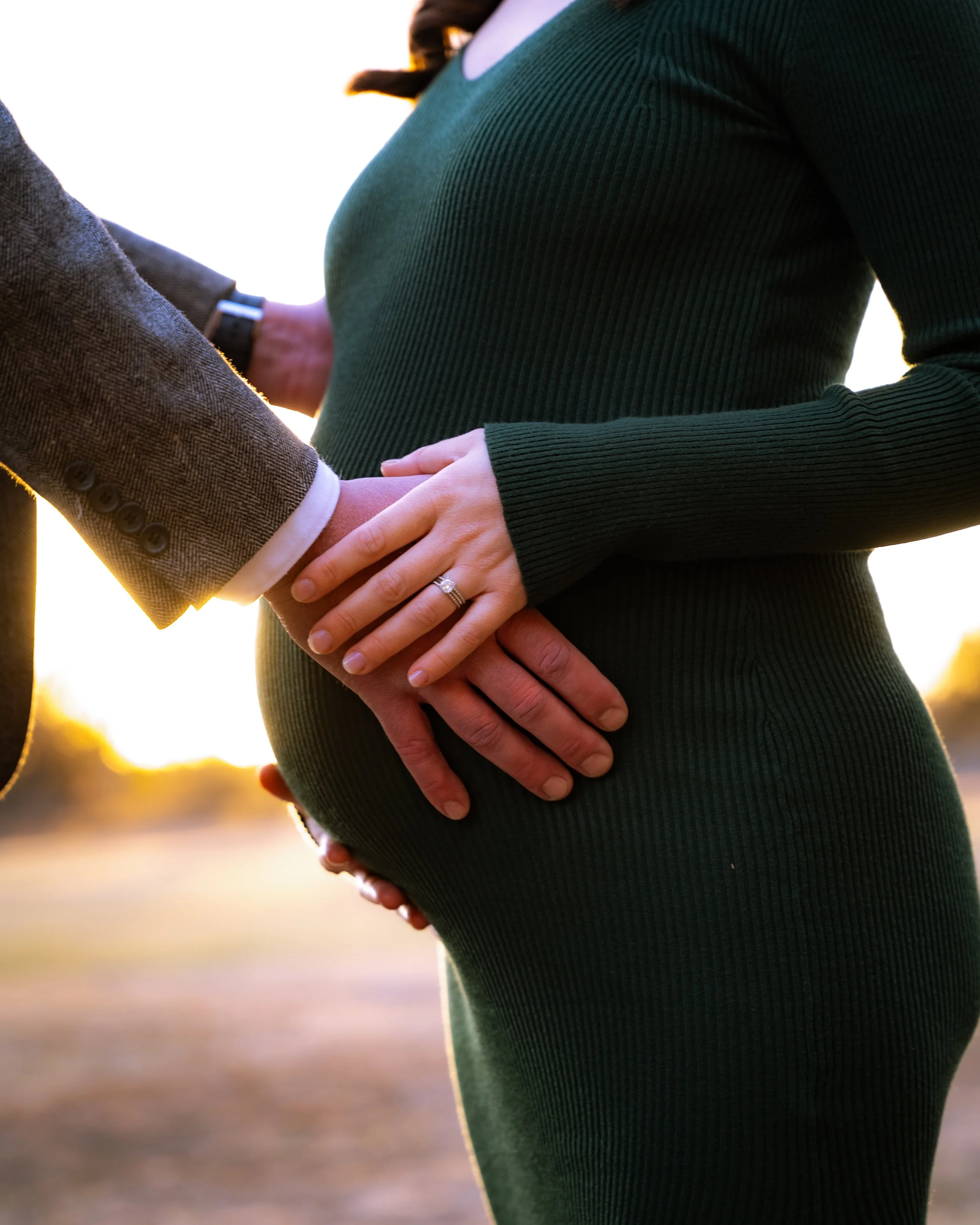 A pregnant woman in a green fitted dress with her partner, holding her belly with both hands, during sunset.
