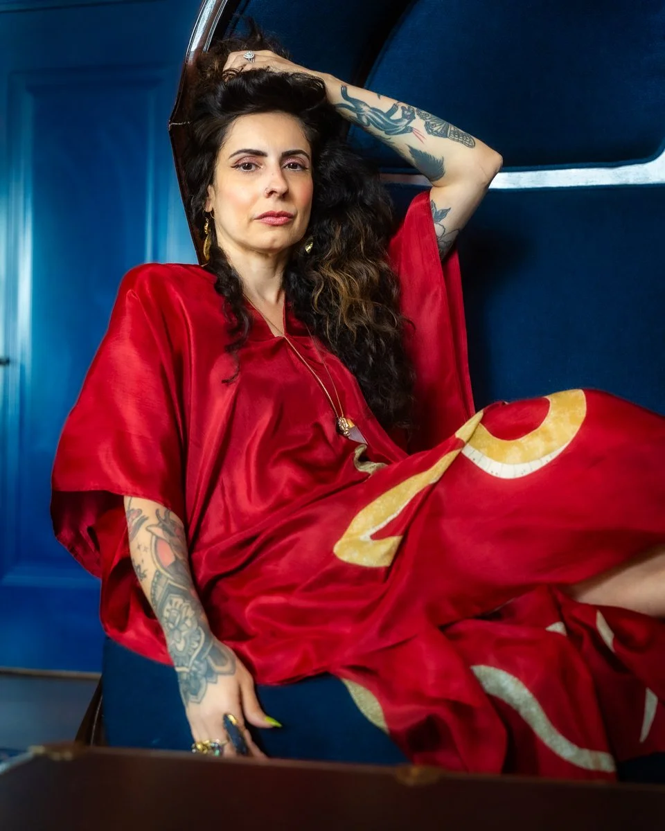 A woman with tattoos and long dark wavy hair sitting on a blue seat, wearing a red satin dress, with her hand on her head and looking at the camera.