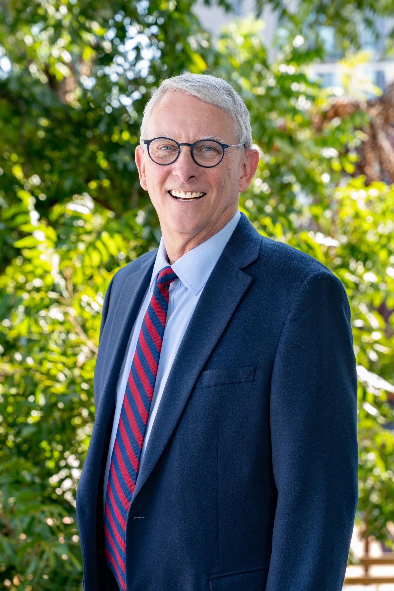 A man with gray hair, glasses, and a suit standing outdoors in front of green foliage, smiling at the camera.