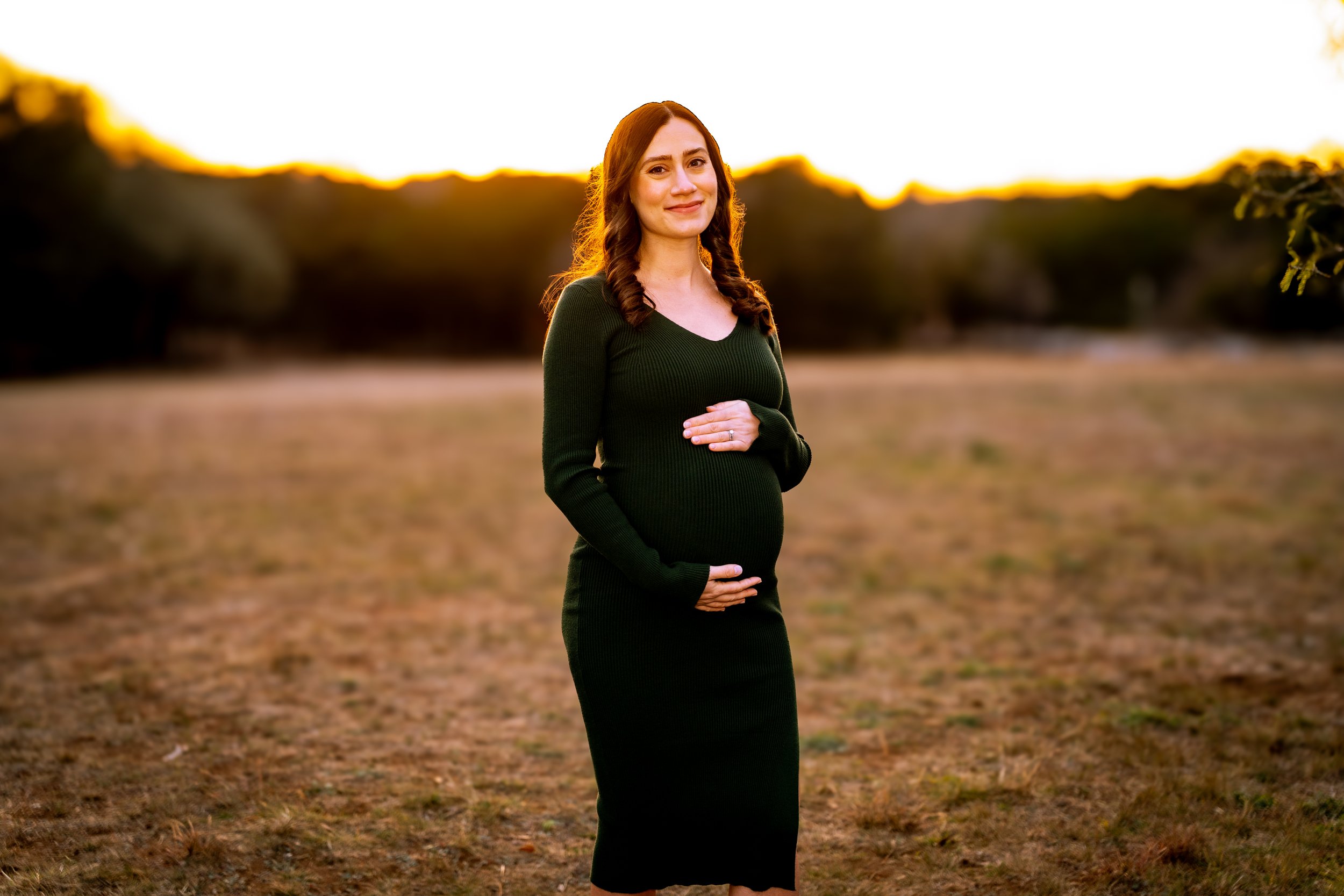 A pregnant woman standing outdoors at sunset, wearing a dark green dress, holding her belly with one hand and resting her other hand on her chest, smiling.
