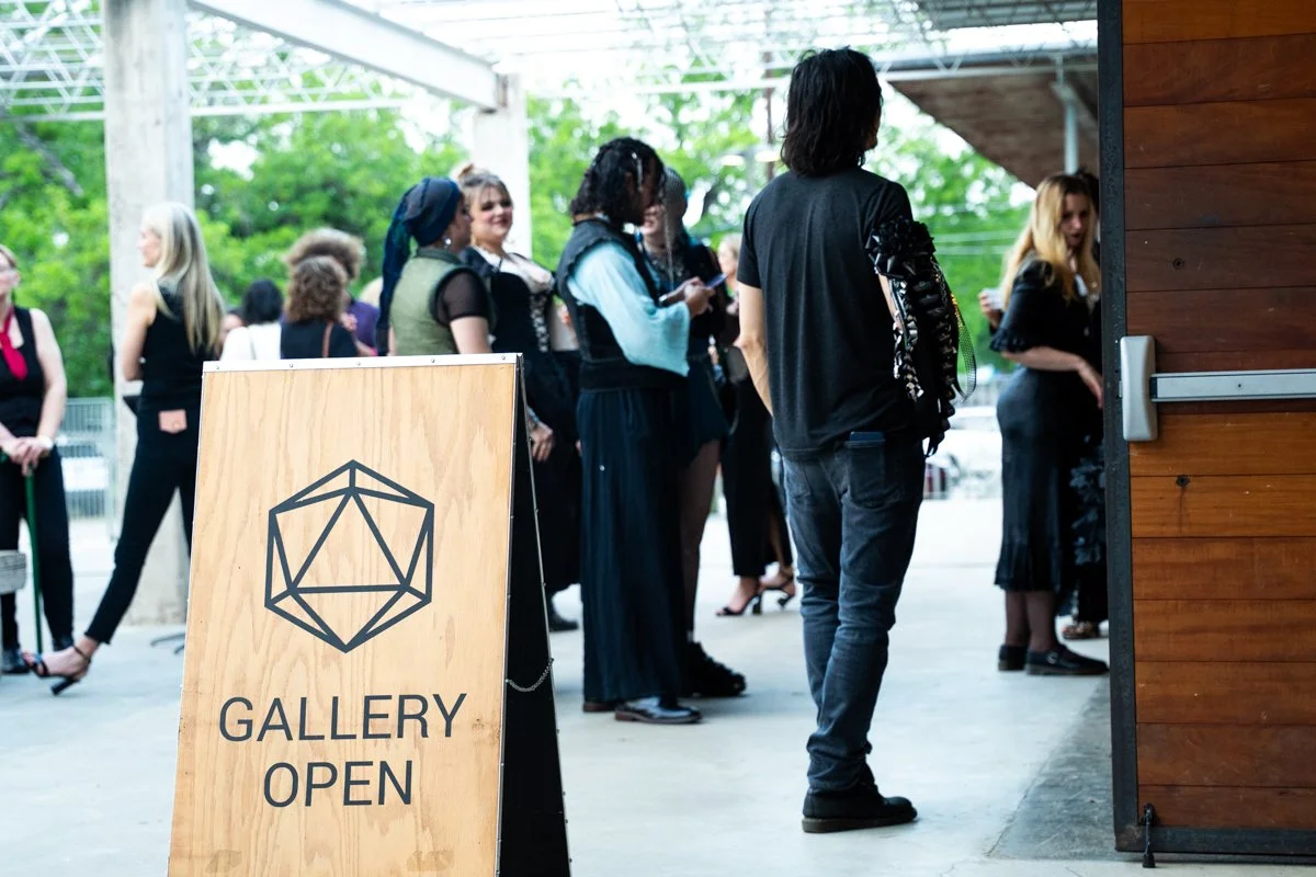 People standing in line outside an art gallery with a wooden sign that says "GALLERY OPEN" and a geometric logo.