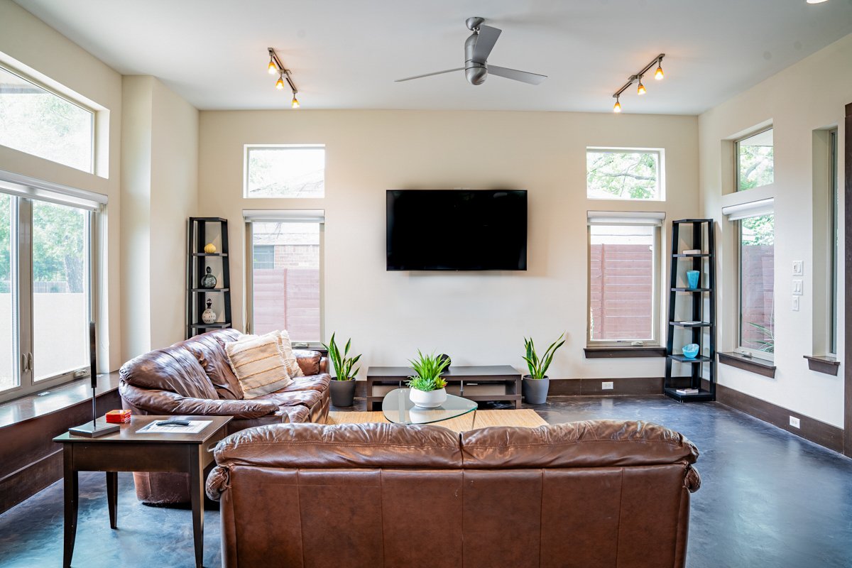 Living room with two brown leather sofas, a glass coffee table, three black shelves with decorations, a wall-mounted TV, four large windows, and a ceiling fan.