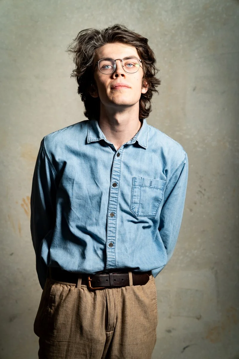 Young man with wavy brown hair and glasses, wearing a light blue button-up shirt and beige pants, standing against a textured neutral background.