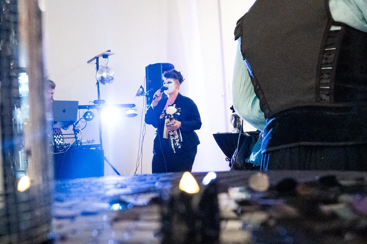 A woman singing into a microphone at an indoor event, holding a bouquet of flowers, with DJ equipment and a disco ball in the background.