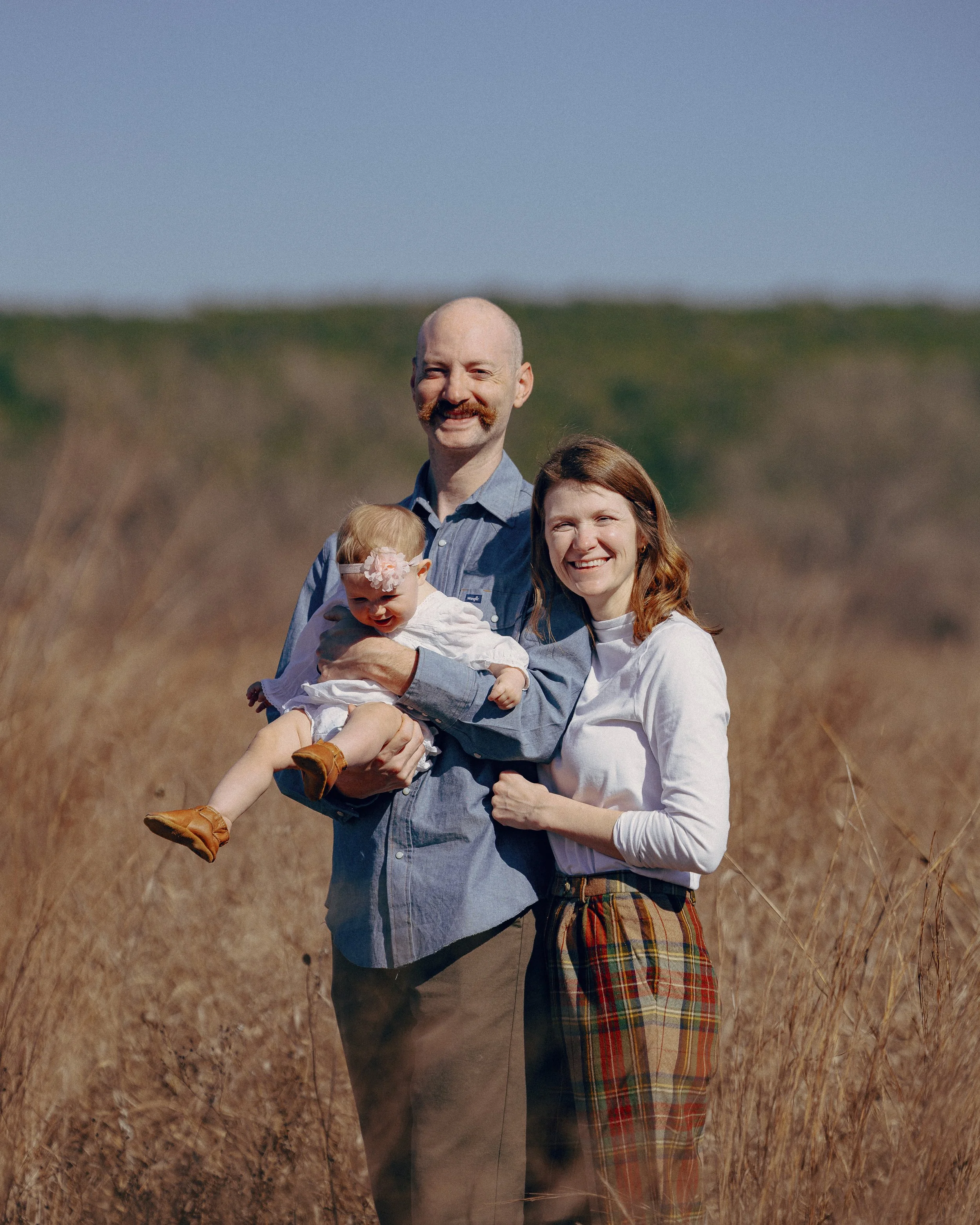 A family of three standing in a field of tall grass on a sunny day, with a man holding a young girl and a woman smiling beside them.