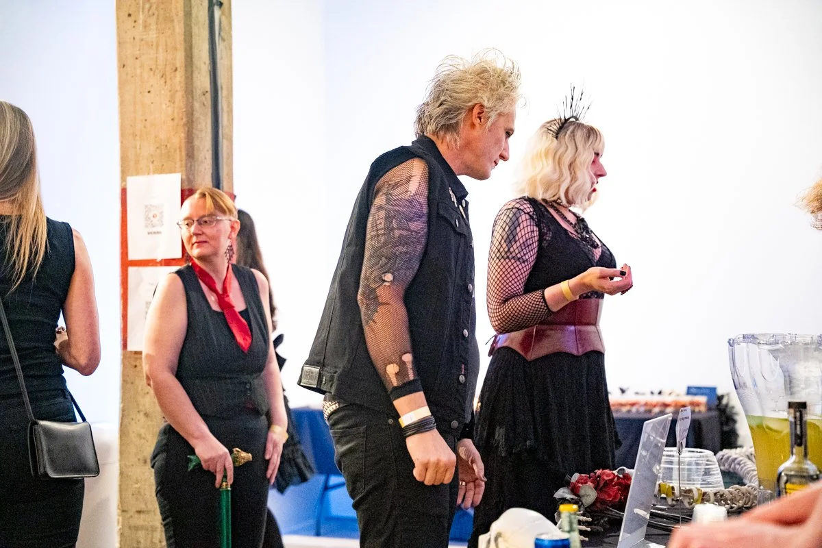 People at an event, interesting attire, standing near a table with drinks and snacks, some looking at a computer or phone.