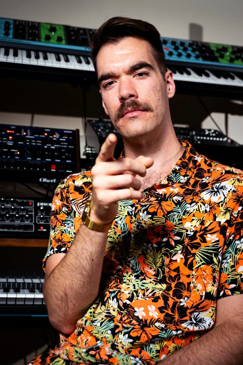 A man with a mustache and dark hair is pointing at the camera. He is wearing a colorful Hawaiian shirt and a gold watch. Behind him is a music studio with multiple synthesizers and electronic music equipment.