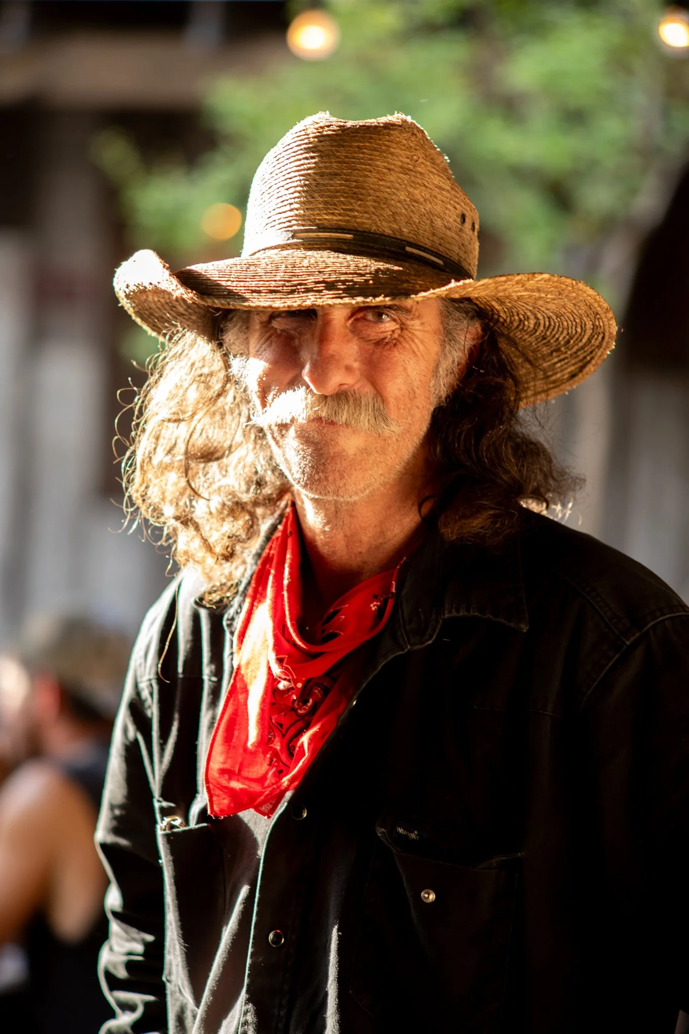 A man with long, curly hair and a mustache, wearing a straw hat and a red bandana around his neck, standing outdoors in sunlight.