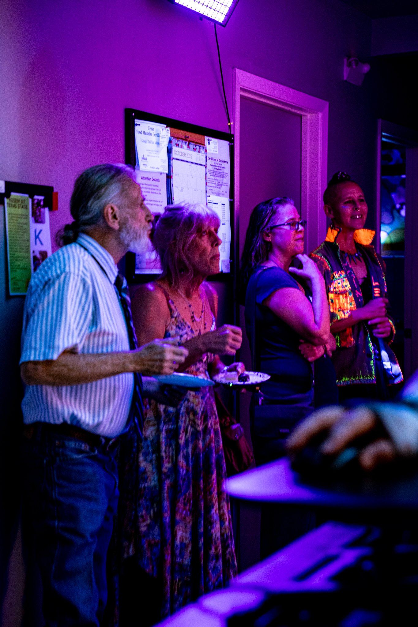 Four people standing in a dimly lit room with purple and blue lighting, some holding plates with food, watching something out of frame. A bulletin board is on the wall behind them.