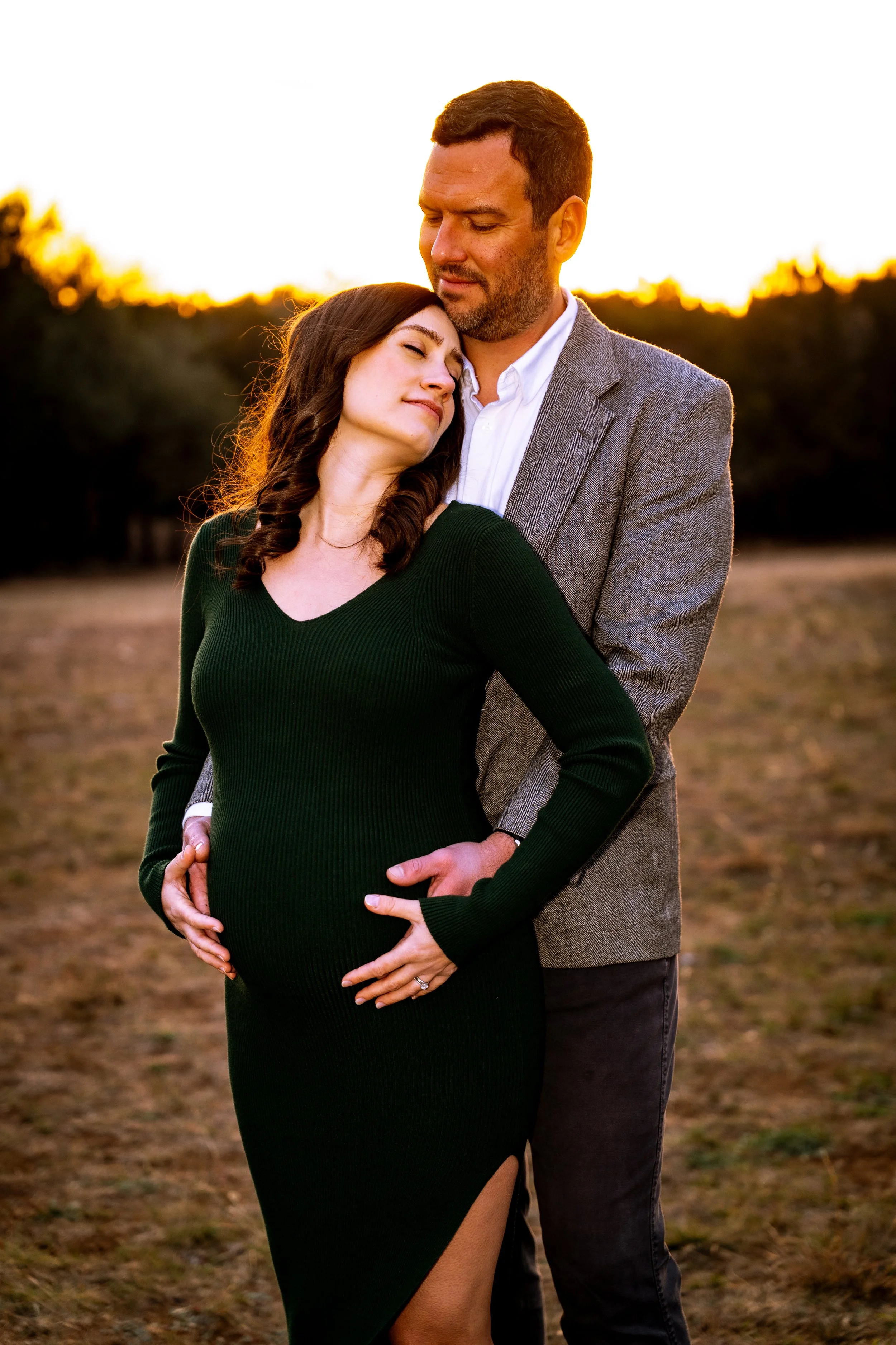A pregnant woman and a man standing outdoors at sunset, with trees in the background. The woman is wearing a black dress with a slit, and the man is dressed in a grey blazer and white shirt. The woman is leaning her head on the man's chest, and the m