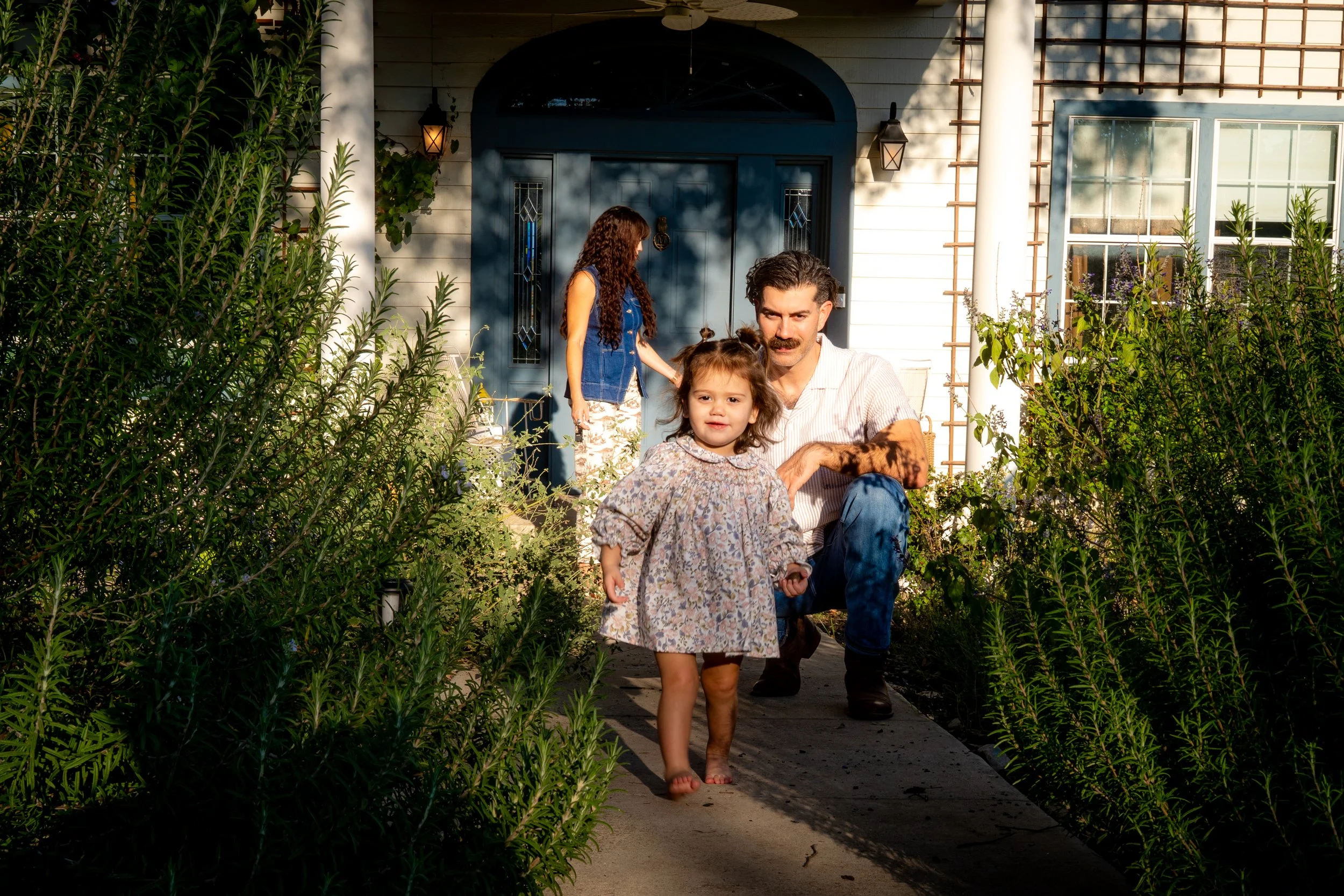 A man crouching on the front walkway of a white house with a dark blue door and a large window, accompanied by two young girls. One girl, barefoot and wearing a floral dress, is walking toward the camera, and the other girl, with long curly hair and 