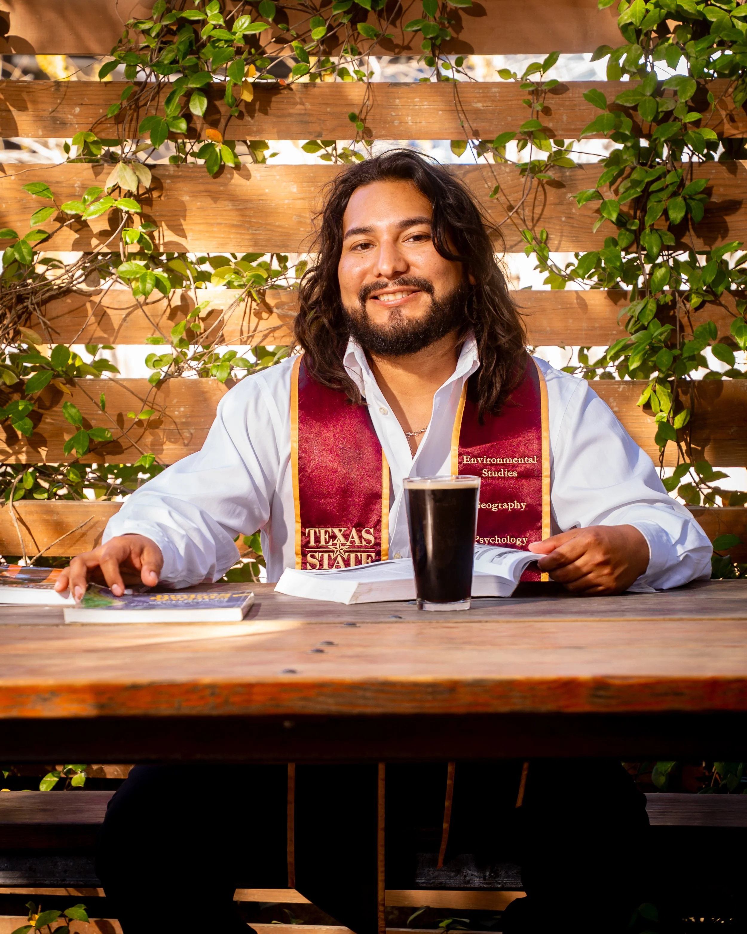 A man with long dark hair and a beard sitting at a wooden table outdoors, smiling at the camera. He is wearing a white shirt and a maroon honors stole, with a drink and some books in front of him, and a backdrop of green foliage and a wooden fence.