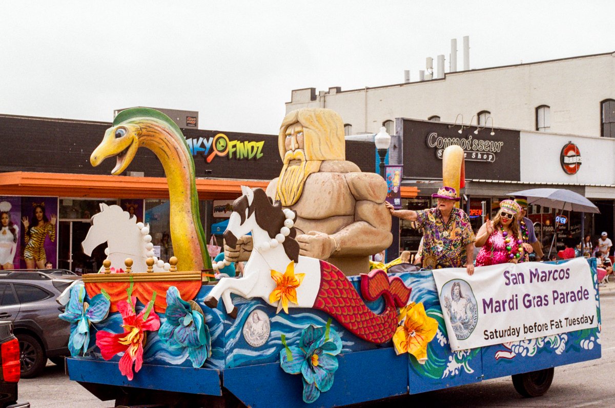 Float in a Mardi Gras parade with a large mermaid, two white horses, a wizard with long blond hair and beard, and tropical flowers, featuring a banner for the San Marcos Mardi Gras Parade scheduled for Saturday before Fat Tuesday.